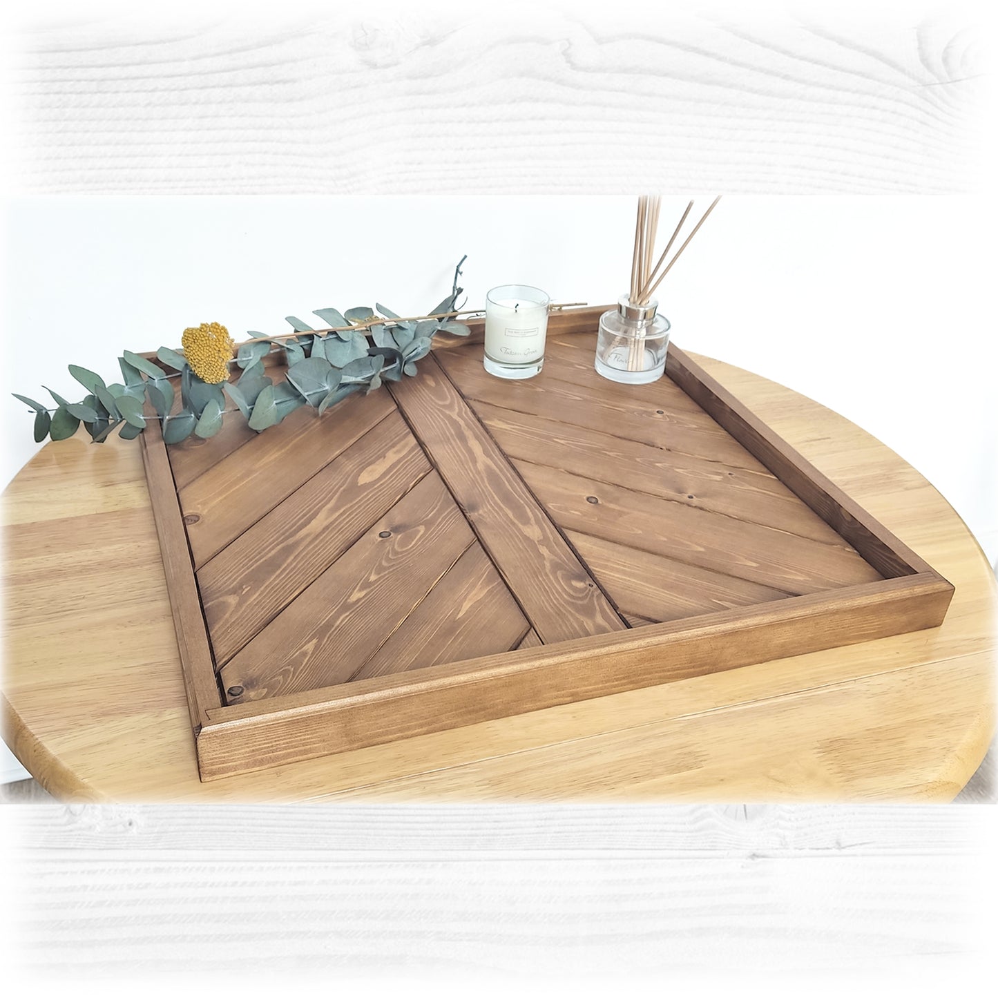 An Ottoman coffee table serving tray in pine, finished in an oak stain and lacquer placed on a wooden table with a white background. Dry flowers, a candle and a diffuser are sitting on top