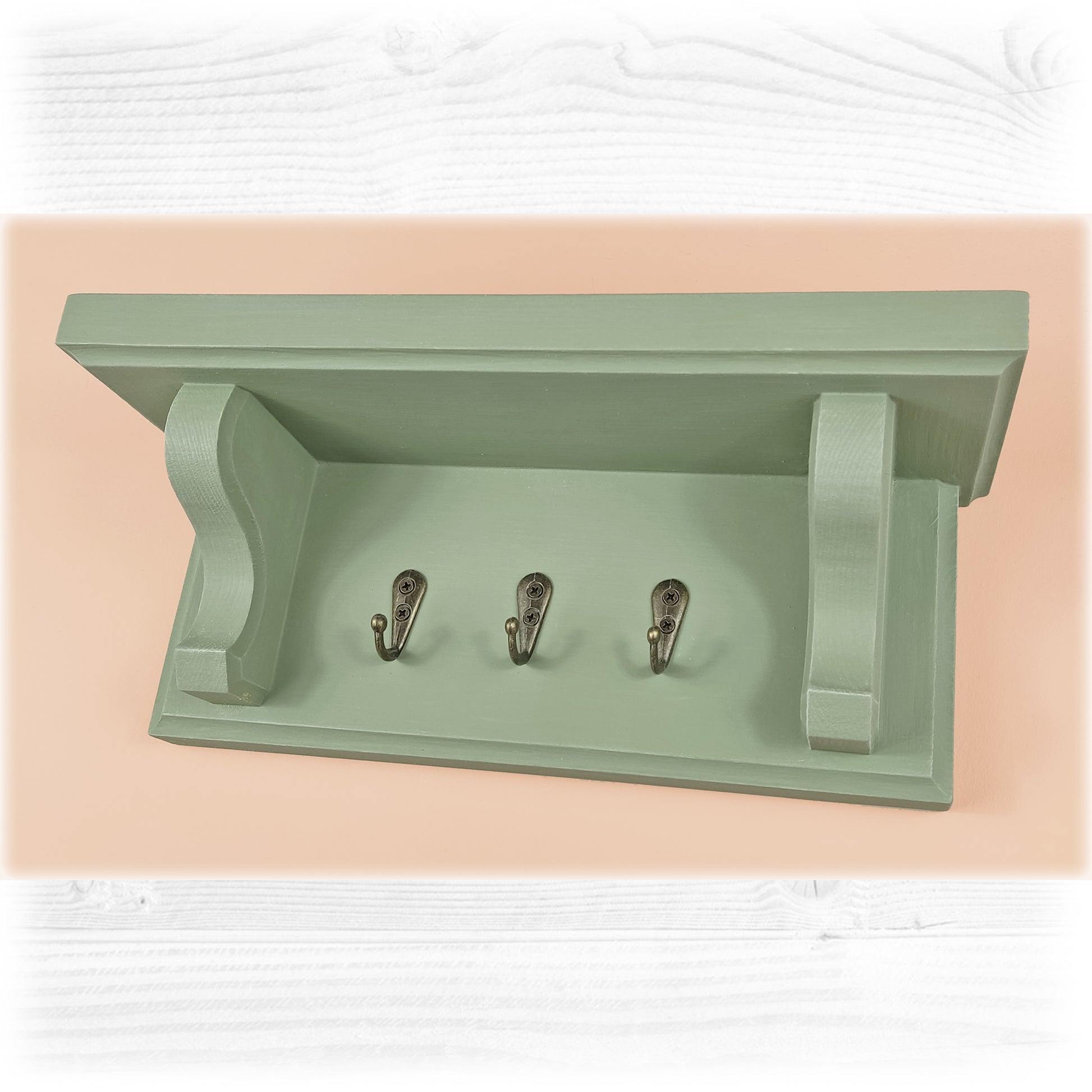 A sage green solid wooden floating key rack with shelf and three bronze key hooks on a pink background