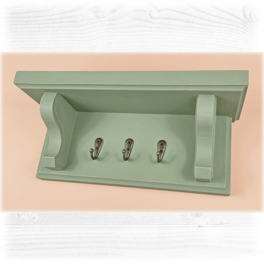 A sage green solid wooden floating key rack with shelf and three bronze key hooks on a pink background
