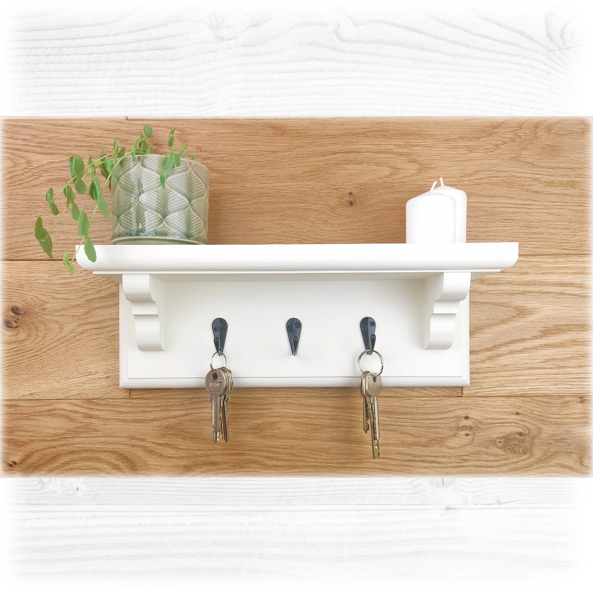 A white wooden floating key rack with shelf and black key hooks on an oak wooden background with a plant and a candle on top and keys hanging