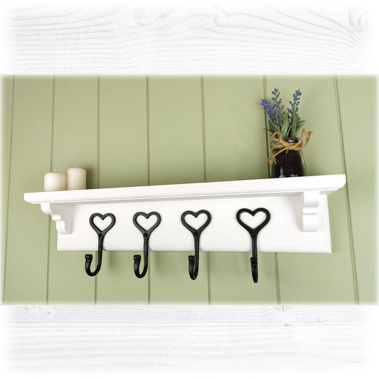 A floating wooden white coat rack with shelf and cast iron black heart shaped coat hooks on a green cladded background