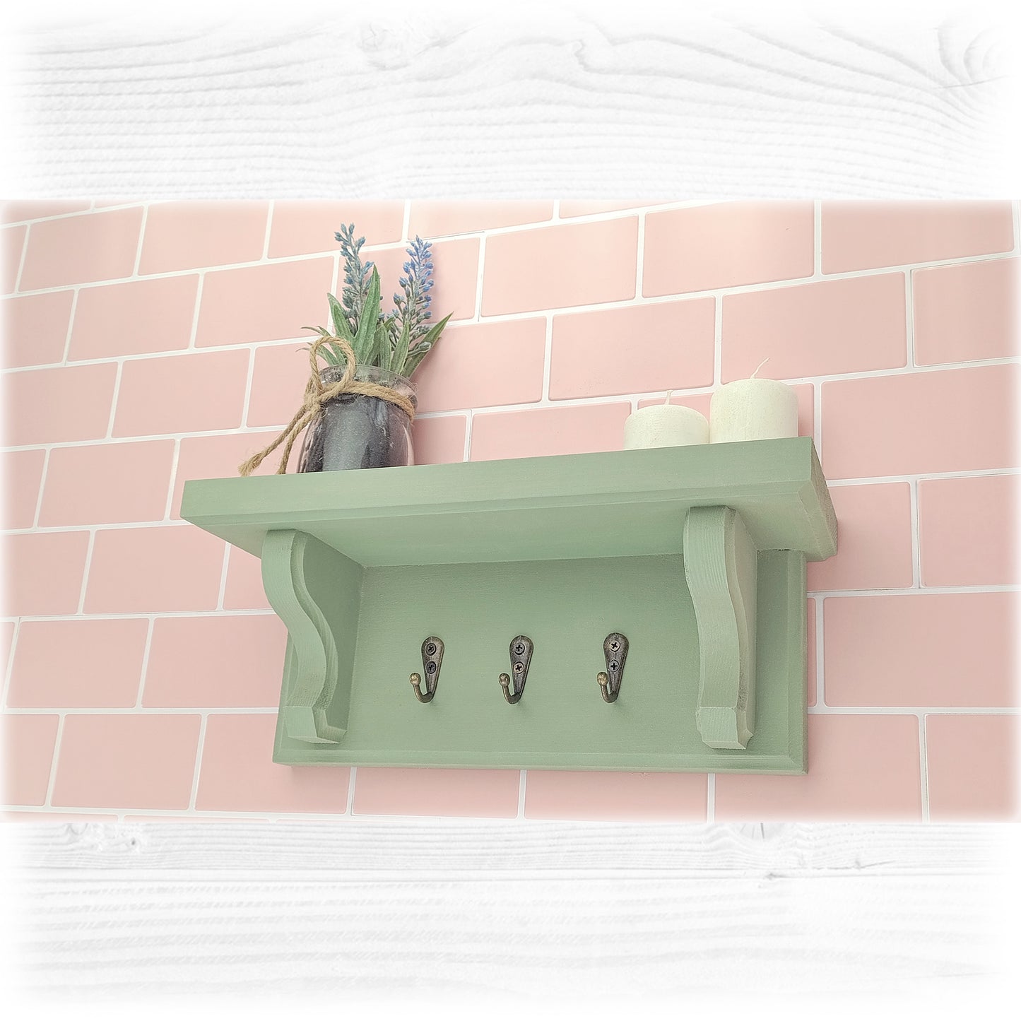 Green key rack with shelf and a small pot of flowers against a pink tiled wall.