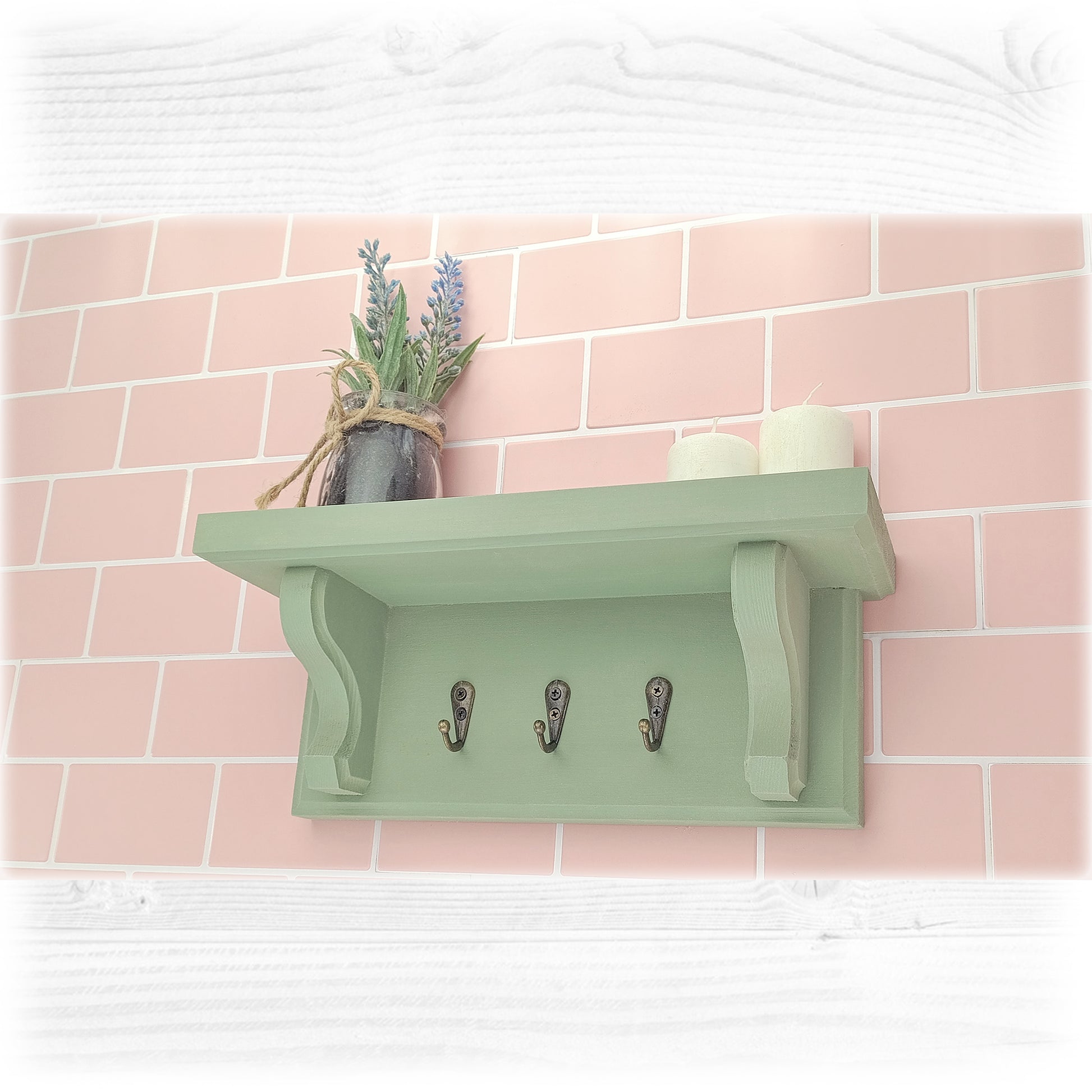 Green key rack with shelf and a small pot of flowers against a pink tiled wall.