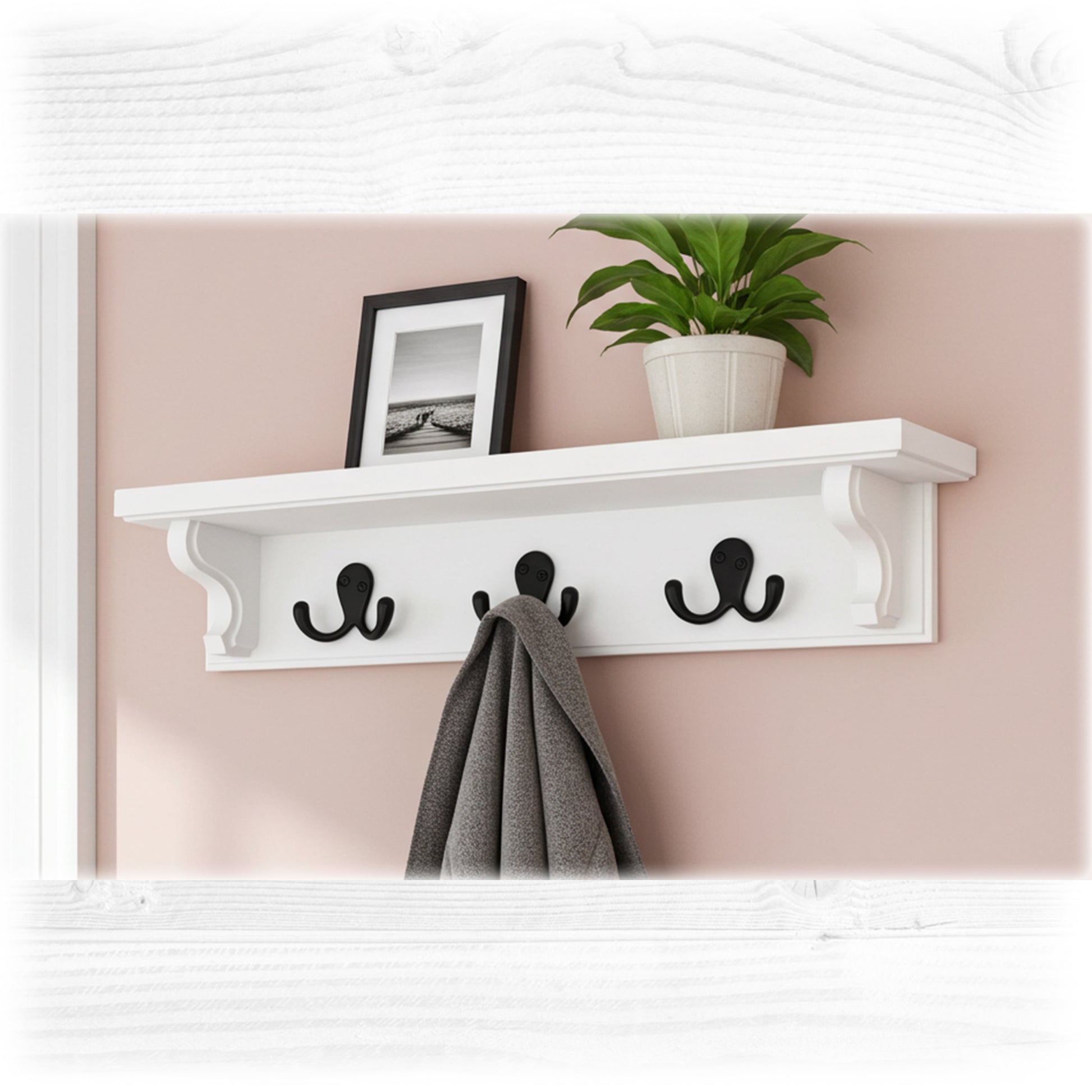 White wall shelf with three black hooks, a plant, and a framed picture on a pink wall.